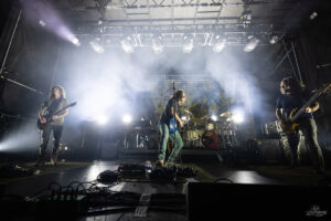 Rock band on stage performing at night with lead singer/guitarist center and two guitarists on sides, bright stage lights and a palm-themed backdrop behind the drums.