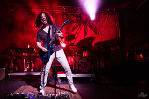 Guitarist with curly hair playing a blue electric guitar on stage under red lights, front and center.