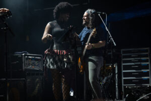 Two musicians on a dimly lit stage share a laugh during a live performance: a woman in a black top and plaid skirt beside a male bassist with long hair and headband.