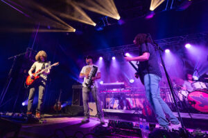 Four musicians perform on a purple-lit stage: guitarist, keyboardist, bassist, and drummer in the background under vibrant lights that sweep across the stage.