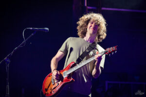 Guitarist with curly hair playing a red electric guitar on stage, microphone nearby.