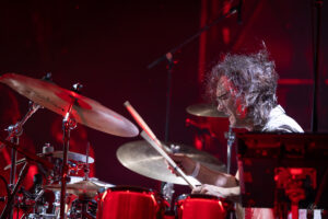 Drummer mid-performance behind a red-lit drum kit with sticks raised and a focused expression on stage.