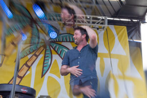 Man on stage smiling, hand on head, wearing a blue short-sleeve shirt, with a yellow palm-tree backdrop behind him.
