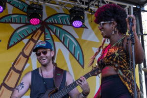 Two musicians on a sunny stage: a smiling male bass guitarist in sunglasses and cap, and a female singer in a tie-dye top with a microphone beside her on stage backdrop with palm trees.