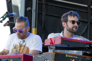 Two male musicians wearing sunglasses playing red keyboards on a stage set-up, with one singing into a mic nearby.