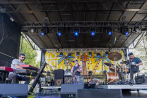 Outdoor stage with a three-person band performing: keyboardist on the left, bassist center, drummer on the right; blue stage lights and a tropical yellow backdrop with palm trees behind them.