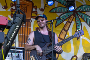 Musician wearing sunglasses and a blue cap plays a brown bass guitar on a sunny stage with a yellow palm mural backdrop and stage lights above.