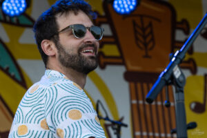 Male singer with sunglasses and a patterned shirt performing on a stage beside a microphone.