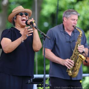 Singer wearing hat and sunglasses belts out a note into a microphone; saxophonist in background performs in a park setting.