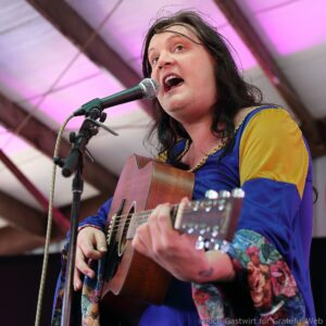 Female vocalist singing into a microphone while strumming an acoustic guitar on stage, wearing a blue and yellow outfit with floral sleeves.
