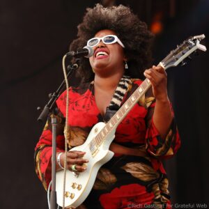 Female guitarist singing into a microphone, wearing white sunglasses and a red patterned outfit on stage.