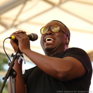 Singer wearing yellow-tinted sunglasses sings into a handheld microphone at close range on stage.