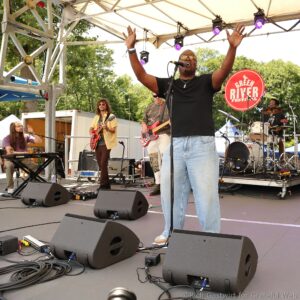 Male singer in black shirt and sunglasses on an outdoor stage, arms raised, with band members and instruments behind him; Green River Festival sign visible in background.