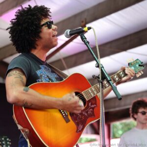Male guitarist singing into a microphone on stage, sunburst acoustic guitar visible in foreground, wearing sunglasses.