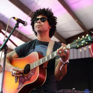 Male guitarist with curly hair and sunglasses plays a sunburst acoustic-electric guitar on stage.