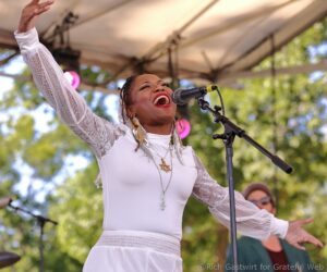 Female gospel singer performing on an outdoor stage, arms raised, singing into a microphone in a white lace outfit.