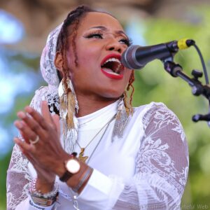 Close-up of a female singer outdoors, singing into a microphone while wearing a lace headscarf and bold red lipstick close to camera.