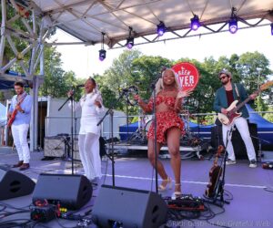 Four-member band performs on an outdoor festival stage; two women sing center while a guitarist and a bassist play nearby under purple lights.