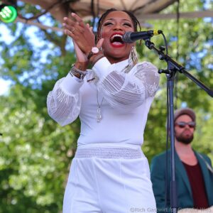 Female singer in a white outfit performing on an outdoor stage with a microphone.