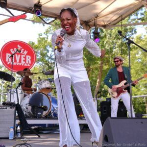 Female singer in white performs into a microphone on an outdoor festival stage, with a band in the background and a red festival sign nearby.