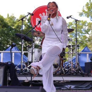 Female singer in white outfit performs on an outdoor stage, holding a microphone with drums in the background.