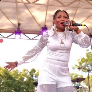 Female singer in a white lace outfit performs on an outdoor stage, gripping a microphone with purple stage lights in the background.