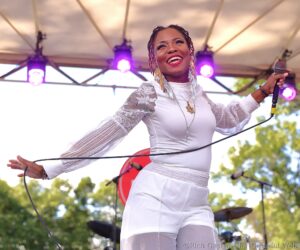 Female singer on stage smiling, holding a microphone, wearing white lace outfit and gold jewelry with purple stage lights behind her.