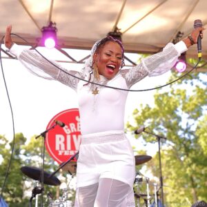 Female singer on stage with arms raised, wearing a white lace outfit and singing into a microphone under pink stage lights.