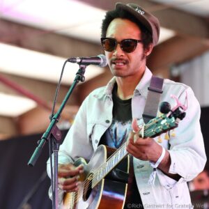Male guitarist wearing sunglasses and a hat sings into a microphone while playing an acoustic guitar on stage