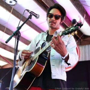 Musician wearing sunglasses and a hat, singing into a microphone while playing an acoustic guitar on stage.
