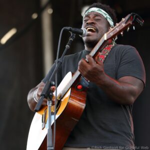 Male guitarist singing into a microphone while playing an acoustic guitar on stage, wearing a green patterned headband and black shirt.