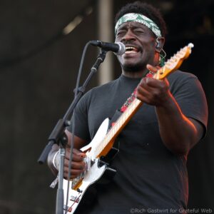 Male guitarist and vocalist wearing a green patterned headband, singing into a microphone while playing a white electric guitar on stage.
