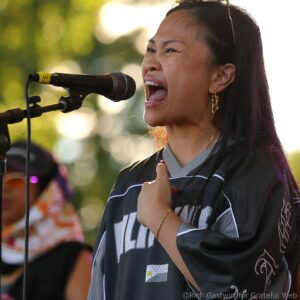 Woman singing into a handheld microphone during an outdoor performance, wearing dark clothing and earrings.