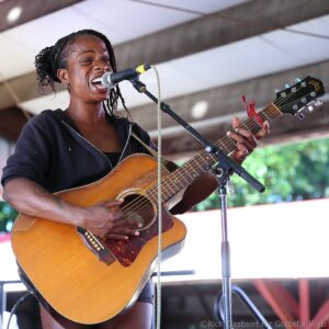 Female musician singing into a microphone while playing an acoustic guitar on stage.