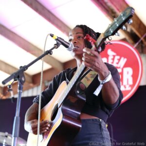Black female musician with braided hair playing an acoustic guitar and singing into a microphone on stage.