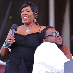 Woman singing into a microphone on stage, wearing a black dress and turquoise jewelry, with another person in white beside her behind.