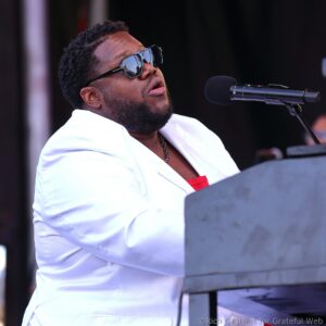 Male singer wearing a white blazer and sunglasses sings into a microphone on stage.