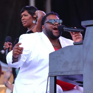 Male singer in a white blazer and sunglasses sings into a microphone on stage, with a female backup singer blurred in the background.