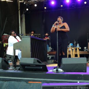 Female singer in a black dress performing on an outdoor stage with a pianist in a white suit and other musicians in the background.