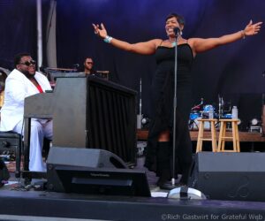 Stage performance: female singer in a black dress with arms outstretched at a microphone, while a pianist in a white suit plays nearby on a dark stage.