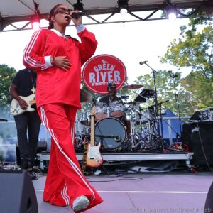 Singer in a red tracksuit performing on stage, microphone in hand, with a guitar and drum set visible behind and a Green River Festival sign in the background.