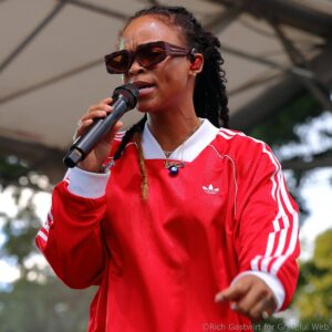 Singer performing into a handheld microphone, wearing a red Adidas jacket and brown sunglasses with braided hair visible in the background, outdoors.