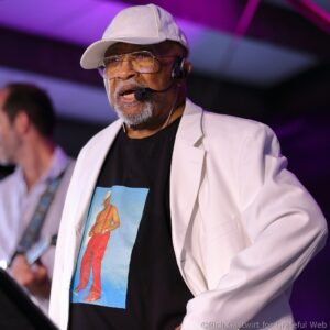 Man performing on stage with a white cap, glasses, and headset microphone under purple lighting.