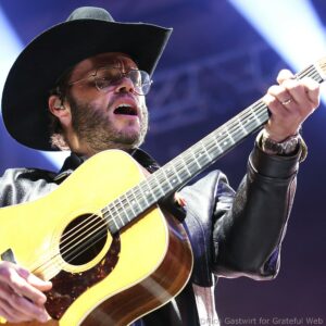 Close-up of a guitarist wearing a black hat and glasses, singing into a mic while strumming an acoustic guitar onstage.