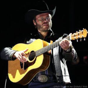 Male musician in a black cowboy hat and glasses playing an acoustic guitar on stage.