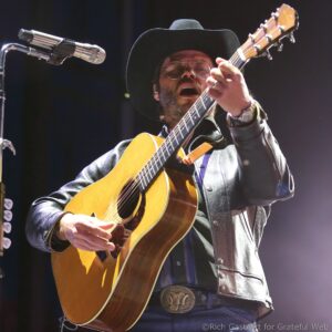 Male musician wearing a black hat and sunglasses, singing into a microphone while playing an acoustic guitar on stage.