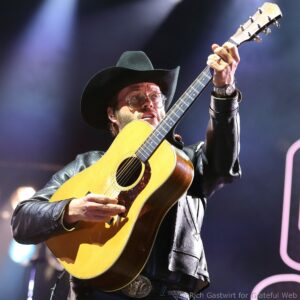 Guitarist wearing a black cowboy hat and glasses plays an acoustic guitar on stage during a live performance.