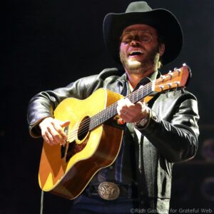 Male musician wearing a black cowboy hat and glasses, strumming an acoustic guitar on stage.
