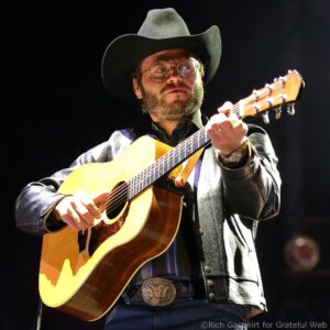 Male musician wearing a cowboy hat and glasses, playing an acoustic guitar on stage.