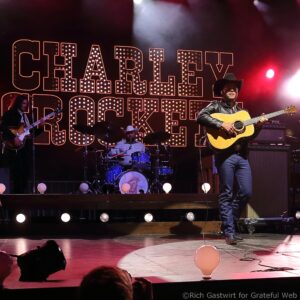 Guitarist in a cowboy hat and leather jacket plays an acoustic guitar on a dim stage with a dotted-backlit sign and balloons on the floor behind him, during a live performance.
